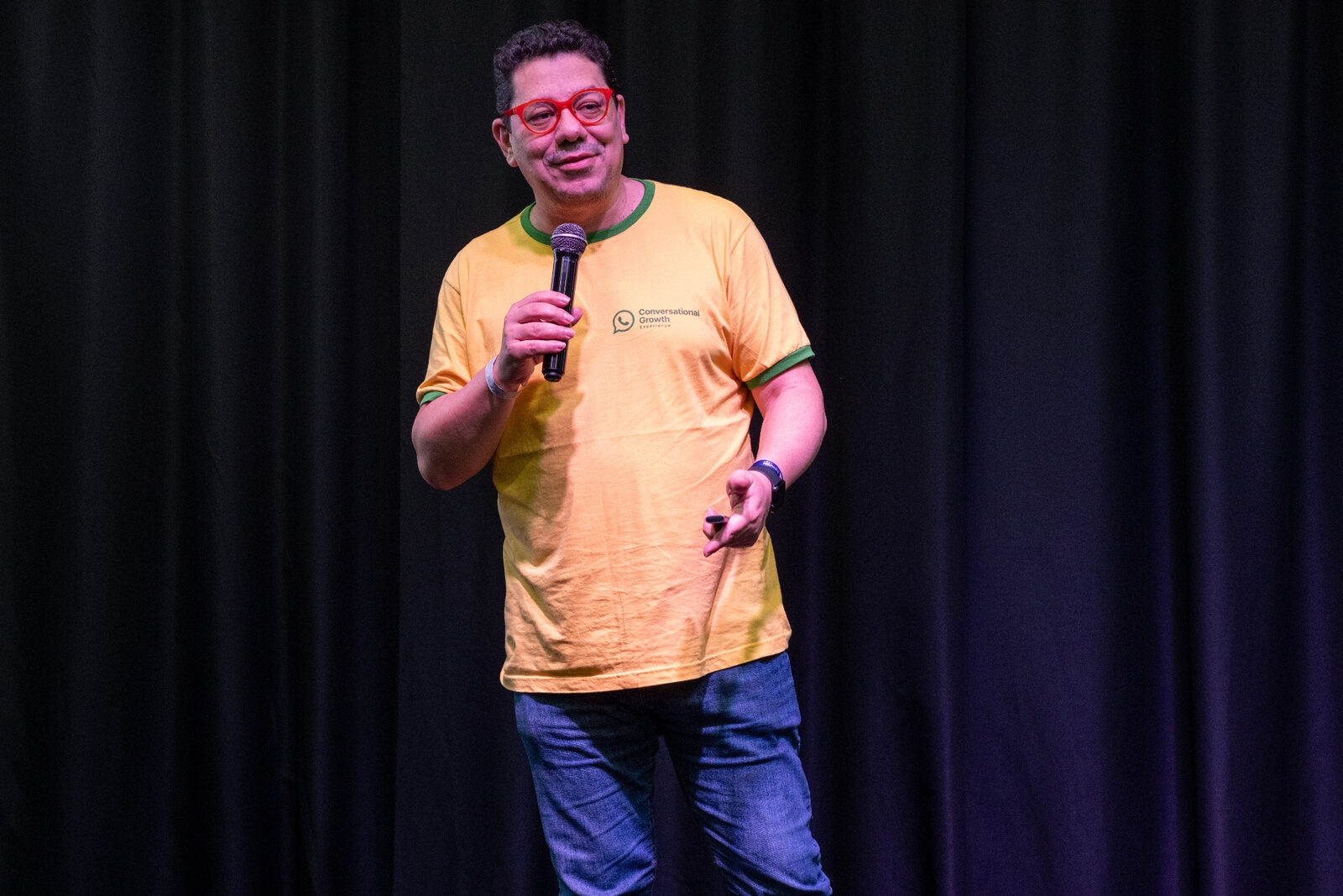Marcus Barboza sur scène, avec microphone, t-shirt orange Conversational Growth Experience et lunettes rouges, présentant la méthodologie MCI